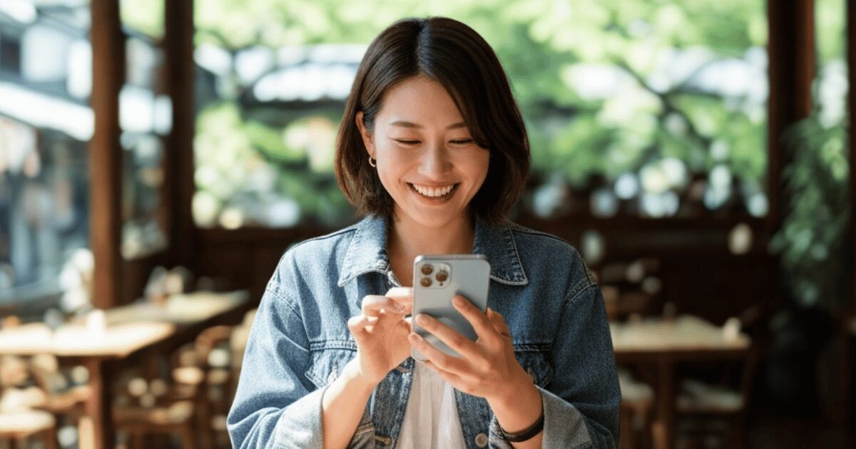 カフェでスマートフォンを操作しながら笑顔を見せる女性。木の温もりと緑に囲まれた落ち着いた空間。