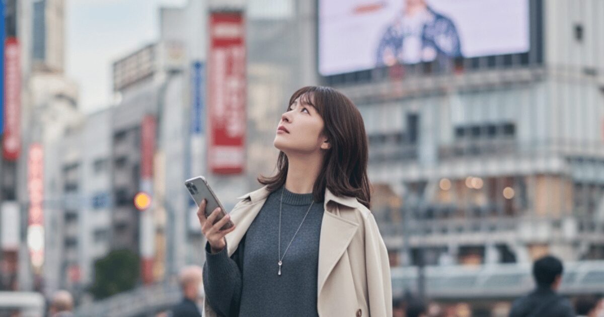スマートフォンを手に街中を歩く女性。高層ビルやデジタルサイネージが並ぶ都会の交差点で空を見上げている。